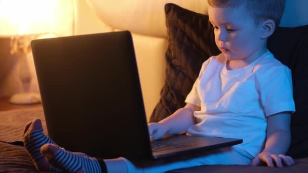 可爱的金发白人男孩深夜独自躺在卧室里，躺在床上，在笔记本电脑键盘上打字，上网或上网。房间里的夜灯，窗外的月光。多莉通过视频滑行. — 图库视频影像