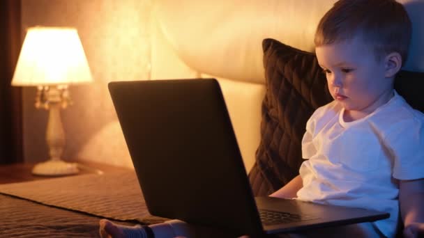 Lindo chico caucásico en el dormitorio tarde en la noche se sienta en la cama, escribiendo en el teclado del ordenador portátil, navegar por Internet y cierra el portátil. luz nocturna en la habitación, luz de luna desde la ventana. Dolly se desliza a través de vídeo . — Vídeo de stock