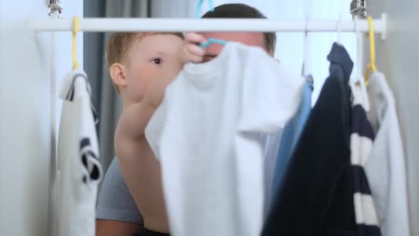 Beyaz erkek baba kollarında çıplak gövdesi olan bir çocuk tutuyor, parlak bir odada dolaptaki askıya beyaz tişört asıyorlar, çocuk kıyafetlerini seçiyorlar. Kamera Dolabın İçinde — Stok video