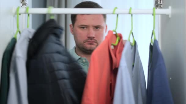 Небритый кавказский мужчина открывает гардероб по утрам и задыхается от одежды, заходит в гардероб, выбирает синюю куртку, чтобы идти на работу. Парень одевается на свидание, камера внутри кабинета — стоковое видео