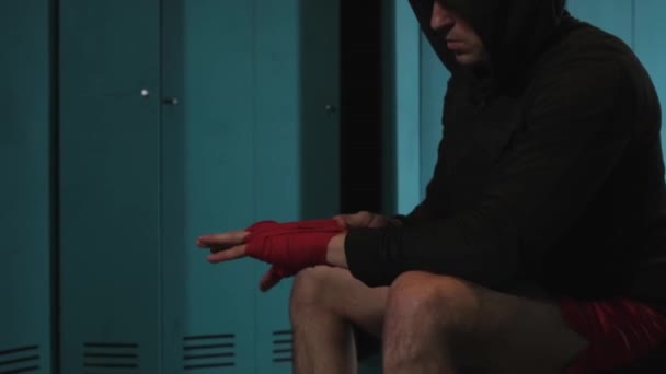 男子拳击手在比赛前用红色绷带捆住双手，一名运动员坐在更衣室里准备参加拳击赛。强壮的运动员在黑暗的体育馆。体育概念和动机. — 图库视频影像