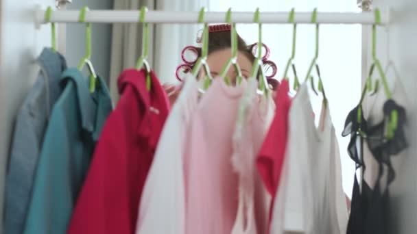 Piękna młoda dziewczyna rano w loki na głowie i w różowym szlafroku otwiera szafę i wybiera biały t-shirt do pracy, przymierza go w pobliżu lustra i zamyka garderobę. — Wideo stockowe