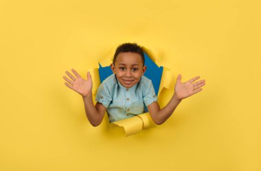 Afro-Amerikalı çocuk, sarı yırtık kağıt arka planda, büyük boy abartılı konsept görüntü üzerinde poz vererek büyük bir şok hissi gösteren ellerini uzatmış kameraya bakıyor..