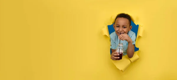 明るく笑顔アフリカ系アメリカ人の少年は 引き裂かれた紙の壁の背景に対して ジュースのボトルを保持し 黒いバーのわらから赤いチェリードリンクを飲みます テキストのためのスペースと健康的なドリンクのコンセプトをデトックス — ストック写真