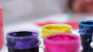 Ressam sarı gouache boya kavanozuna fırça koyar, tablo üzerinde diğer renkli boyalarla birlikte tuval ile birlikte durur. Sanatçı resim çizme sanatı ile meşgul. Yakın çekim videosu