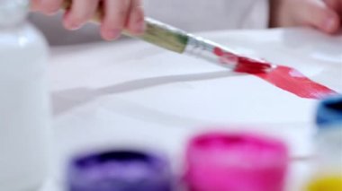 Küçük çocuk arka planda farklı renklerde beyaz kağıda kırmızı boyayla fırça çizer. Sanatçı resim çizme sanatı ile ilgilenir. Videonun yakın çekim görüntüsü