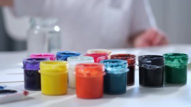 Parlak çocuk odasındaki sanatçı çocuk, beyaz akrilik boyaya daldırıyor ve tuvale çiziyor. Erkek ressam, arka planda küçük kavanozlarda farklı renkte guaj boyaları çiziyor.