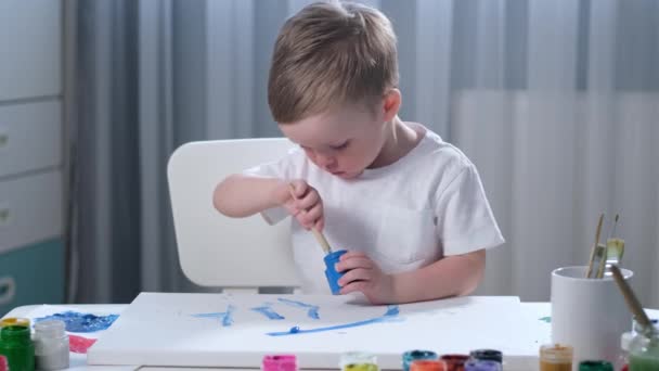 Blond blank jongetje in heldere kinderkamer in wit t-shirt tekent met borstel met blauwe gouache verf op wit doek, maakt penseelstreken tekening beeld. kunstenaar tekent op wit papier. — Stockvideo