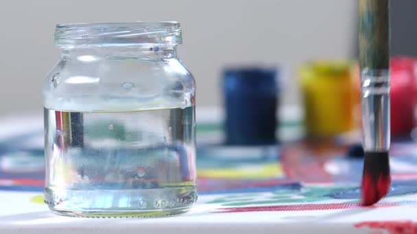 Artist works on picture, draws red straight line on canvas with brush and then washes brush in with jar of clean water. Brush bristles covered with paint are cleaned in close-up. Concept art Studio. — Stock Video