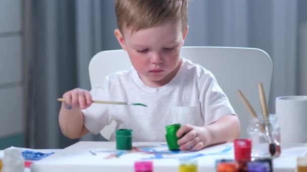 Cute fair-haired Caucasian artist child in white t-shirt with dirty, paint-stained hands, sits at table in childrens room and draws picture with brush with green paint. Boy painter is engaged in art. — Stock Video