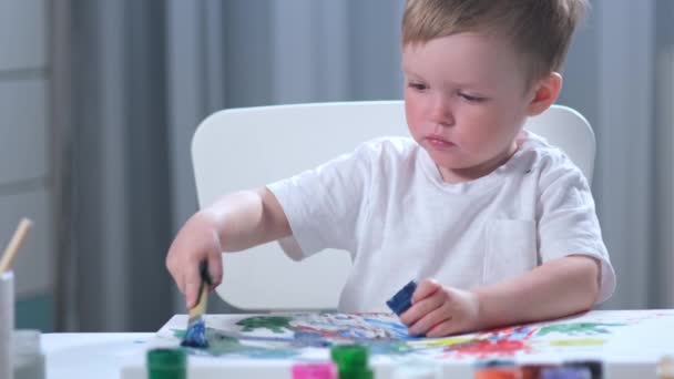 Niño artista caucásico de pelo rubio en camiseta blanca con las manos sucias y manchadas en pintura se sienta a la mesa en la habitación de los niños y dibuja cuadro con pincel con pintura azul. Chico pintor se dedica al arte . — Vídeos de Stock