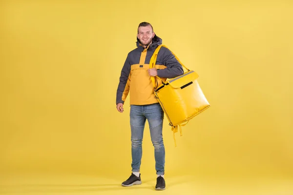 Entrega de comida sonriente joven caucásico en chaqueta amarilla y con bolsa de termo en los hombros sobre fondo amarillo. Concepto de entrega rápida de comida en la ciudad, pancarta con espacio para texto y eslogan — Foto de Stock