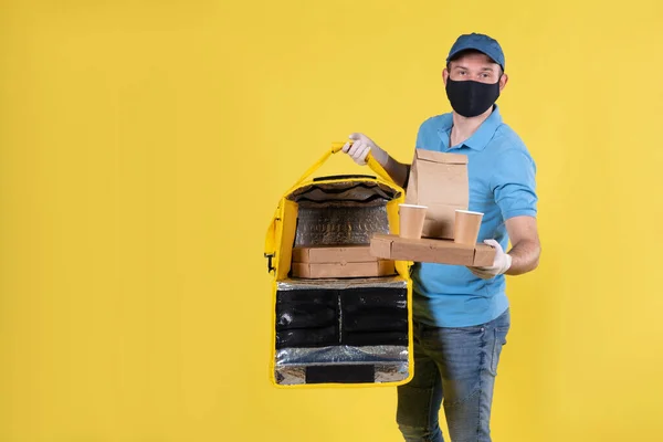 Hombre de entrega de comida para niños con máscara protectora y guantes con Polo azul y gorra mantiene el orden de los clientes en sus manos y bolsa de termo con cajas de pizza y mantiene el orden en el fondo amarillo . — Foto de Stock