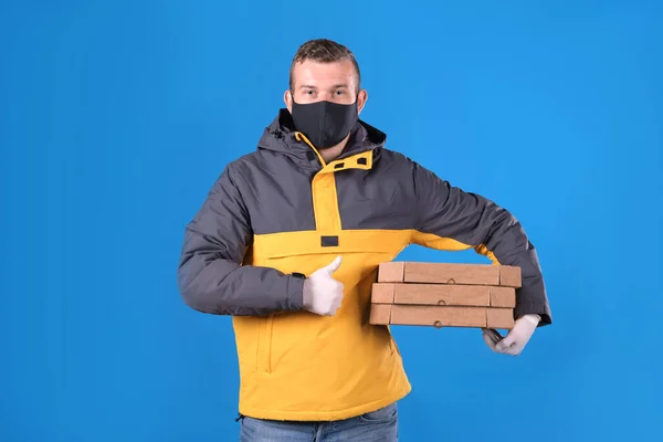 Young cute guy courier in black medical mask and rubber gloves delivering food to your home or office holds an order of pizza in box with an ecological biodegradable packaging on blue background