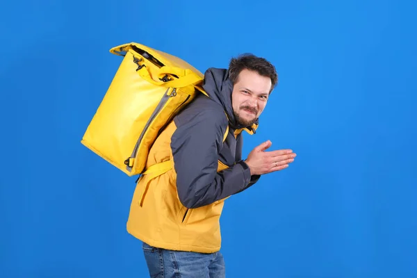 Sly food delivery man with yellow thermos bag is making face and rubbing his hands in anticipation of something on blue fone. Cheerful courier with grin on his face is planning to take over world — Stock Photo, Image