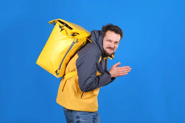 Sly repartidor de comida hombre con bolsa de termo amarillo está haciendo la cara y frotando sus manos en anticipación de algo en fone azul. Un mensajero alegre con una sonrisa en la cara planea apoderarse del mundo — Foto de Stock