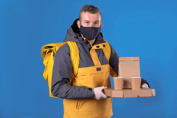 Cute caucasian courier and food delivery man in black mask and rubber gloves in yellow jacket and thermal bag behind his back holds ready order for the client. Food delivery coronavirus pandemic. — Stock Photo, Image