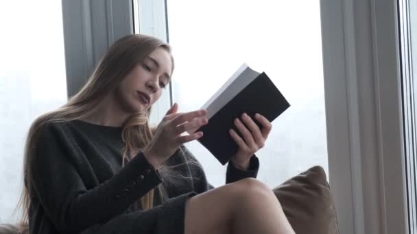 Jovem menina bonita lendo um livro pela janela — Vídeo de Stock