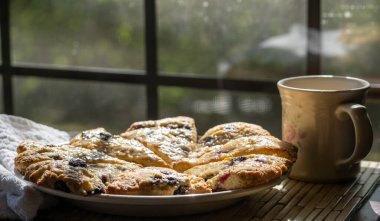 Yaban mersinli çörekler bir fincan sıcak içecekle yuvarlak bir tabakta üçgen şeklinde kesilmiş.. 