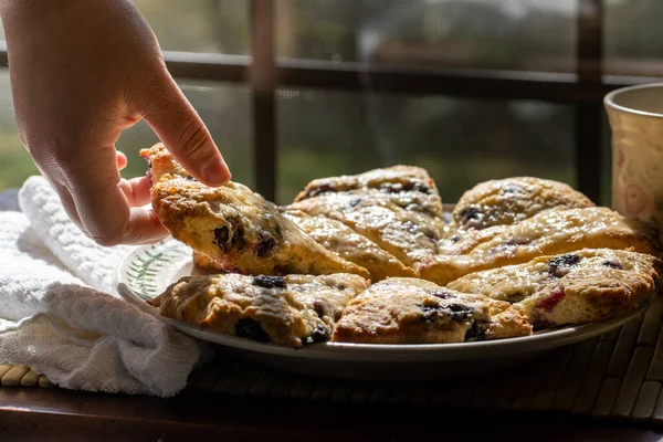 Yaban mersinli çörekler yuvarlak bir tabakta üçgen şeklinde kesilmiş ve bir çörek almak için uzanan bir el var.. — Stok fotoğraf