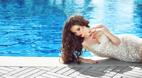 Beautiful woman in Elegant dress. Sexy brunette lying by blue swimming pool. Fashion outdoor portrait. Luxury resort. Cute girl model with red lips makeup and long curly hair. — Stock Photo, Image