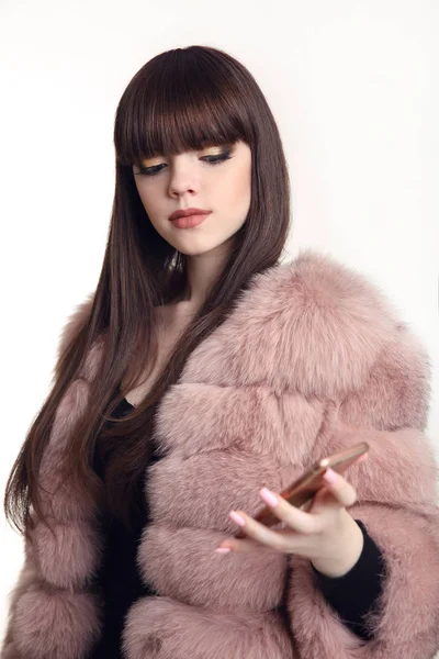 Chica de moda mirada en abrigo de piel rosa sosteniendo teléfono móvil aislado —  Fotos de Stock