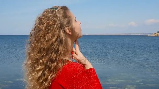 Close up portrait of woman running hand through curly hair blowing in wind by sea on beach. Happy female over blue sky. — Stock Video