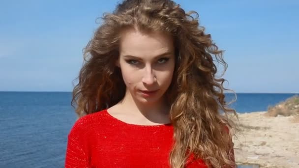 Close up portrait of woman running hand through curly hair blowing in wind by sea on beach. Happy female over blue sky — Stock Video