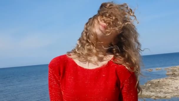 Close up portrait of woman running hand through curly hair blowing in wind by sea on beach. Happy female over blue sky — Stock Video