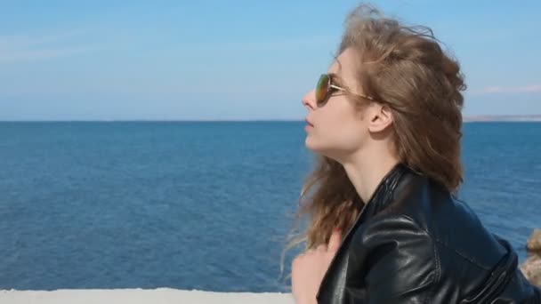 Close up portrait of woman in fashion trendy sunglasses running hand through curly hair blowing in wind by sea on beach. Happy female over blue sky — Stock Video