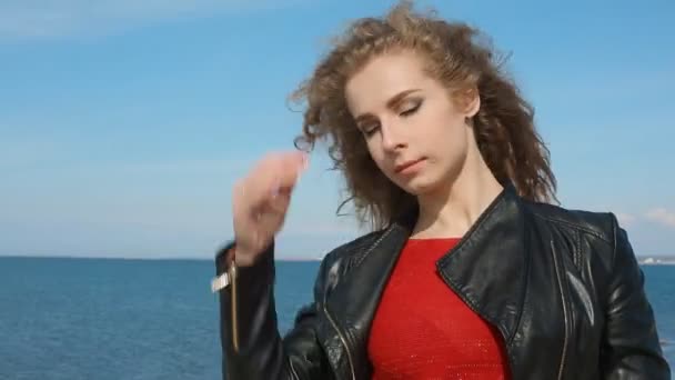 Close-up portret van vrouw opstijgen zwart lederen jas, trendy vrouwen met krullend haar waait in de wind van zee op strand. Gelukkige vrouw over blauwe hemel. — Stockvideo