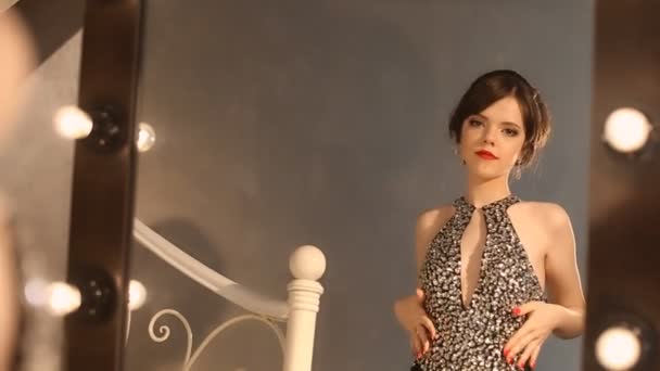 메이크업과 헤어스타일, 매력적인 갈색 빛 전구 드레싱 룸에서 거울 앞에서 포즈와 우아한 검은 드레스에 아름 다운 패션 소녀. — 비디오