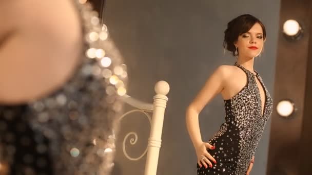 Schöne Mode junge Mädchen in elegantem schwarzen Kleid mit Make-up und Frisur, attraktive Brünette posiert vor dem Spiegel mit Glühbirnen in der Umkleidekabine. — Stockvideo