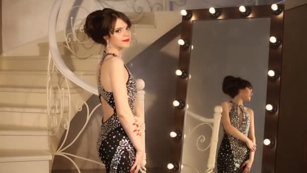 Señora elegante, modelo joven en posando vestido negro de lujo, linda chica delante del espejo con bombillas en el vestidor. Morena con maquillaje y peinado . — Vídeo de stock