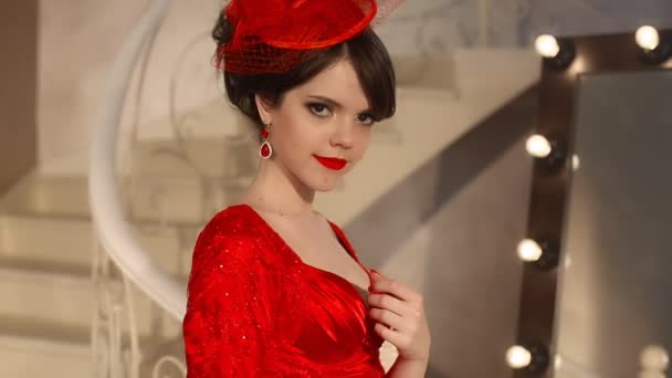 Primer plano de hermosa chica de moda en sombrero retro y vestido rojo, modelo morena con maquillaje de labios rojos, peinado elegante, colgante de las mujeres conjunto de joyas posando por espejo con bombillas para el maquillaje en vestirse — Vídeos de Stock