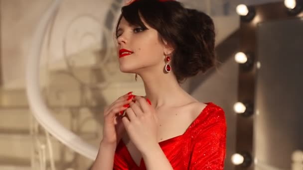 Closeup of beautiful fashion young girl in retro hat and red dress, brunette model with red lips makeup, elegant hairstyle, pendant women set jewelry posing by mirror with bulbs for makeup in dressing — Stock Video