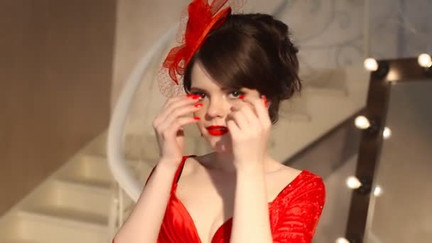 Primer plano de hermosa chica de moda en sombrero retro y vestido rojo, modelo morena con maquillaje de labios rojos, peinado elegante, colgante de las mujeres conjunto de joyas posando por espejo con bombillas para el maquillaje en vestirse — Vídeos de Stock