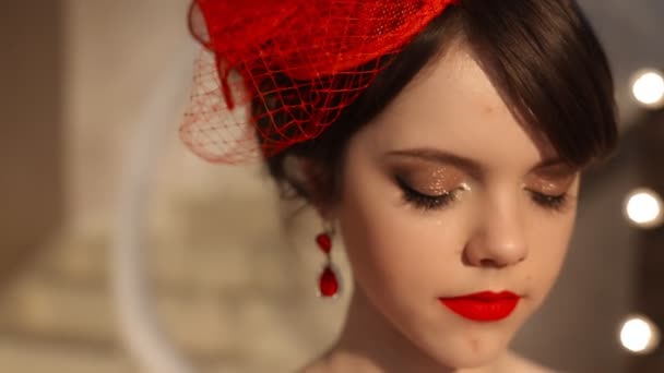 Zbliżenie, piękne Moda dziewczyna w kapeluszu retro i czerwona sukienka, model brunetka z czerwonymi ustami makijaż, elegancką fryzurę, wisiorek kobiety Komplet biżuterii stwarzające przez lustro z żarówki do makijażu w opatrunku — Wideo stockowe