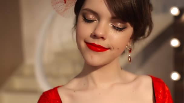 Closeup of beautiful fashion young girl in retro hat and red dress, brunette model with red lips makeup, elegant hairstyle, pendant women set jewelry posing by mirror with bulbs for makeup in dressing — Stock Video