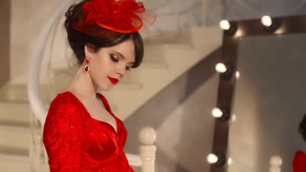 Close-up de bela moda jovem em chapéu retro e vestido vermelho, modelo morena com maquiagem lábios vermelhos, penteado elegante, mulheres pingente definir jóias posando por espelho com bulbos para maquiagem em vestir — Vídeo de Stock