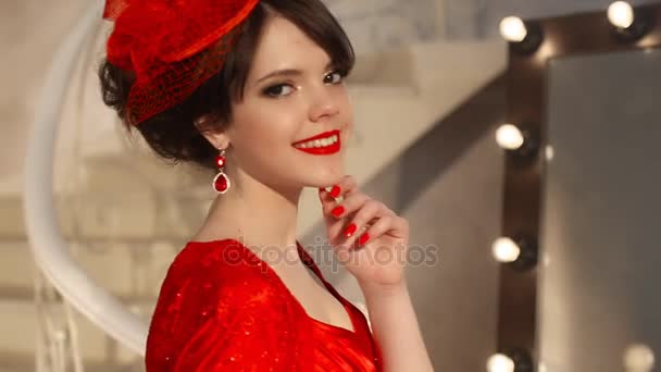 Primer plano de hermosa chica de moda en sombrero retro y vestido rojo, modelo morena con maquillaje de labios rojos, peinado elegante, colgante de las mujeres conjunto de joyas posando por espejo con bombillas para el maquillaje en vestirse — Vídeos de Stock