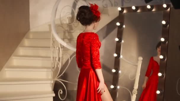 Primer plano de hermosa chica de moda en sombrero retro y vestido rojo, modelo morena con maquillaje de labios rojos, peinado elegante, colgante de las mujeres conjunto de joyas posando por espejo con bombillas para el maquillaje en vestirse — Vídeo de stock