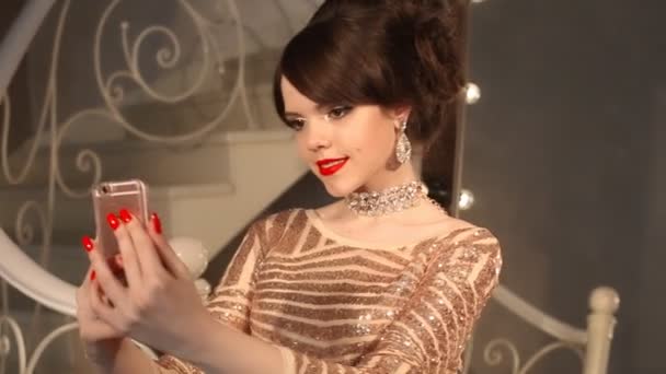 Selfie. Jeune adolescente prenant des photos en robe dorée et bijoux. Élégante dame en robe de paillettes posant par miroir avec des ampoules pour le maquillage, dans un appartement de luxe à la fête . — Video