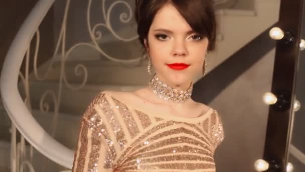 Chica sonriente joven feliz con maquillaje y peinado, joyas de oro, uñas rojas cuidadas. Atractiva joven morena en vestido de lentejuelas posando en la cámara en habitación moderna . — Vídeos de Stock