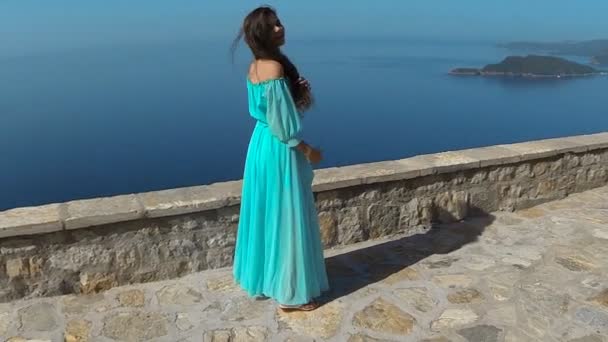 Krásná bruneta usmívající se dívka s zdravé dlouhé vlasy ve větru a šaty. Šťastná mladá žena užívat si přírody a baví nad mořem a modrá obloha. Sveti Stefan, Černá Hora.