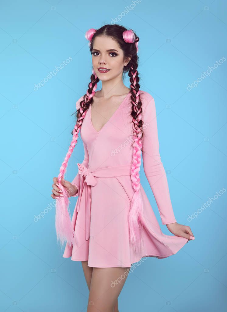 Pretty teen girl with two french braids from pink kanekalon, fashionable hairdo for youth, creative hairdresser beauty salon. Positive brunette posing in cute dress isolated on blue studio background.