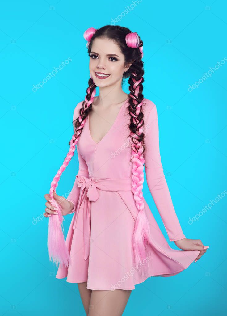 Pretty teen girl with two french braids from pink kanekalon, fashionable hairdo for youth, creative hairdresser beauty salon. Positive brunette posing in cute dress isolated on blue studio background.