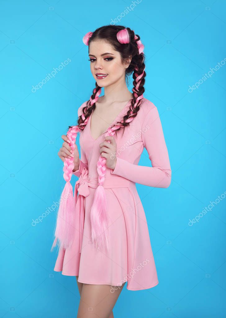 Pretty teen girl with two french braids from pink kanekalon, fashionable hairdo for youth, creative hairdresser beauty salon. Positive brunette posing in cute dress isolated on blue studio background.