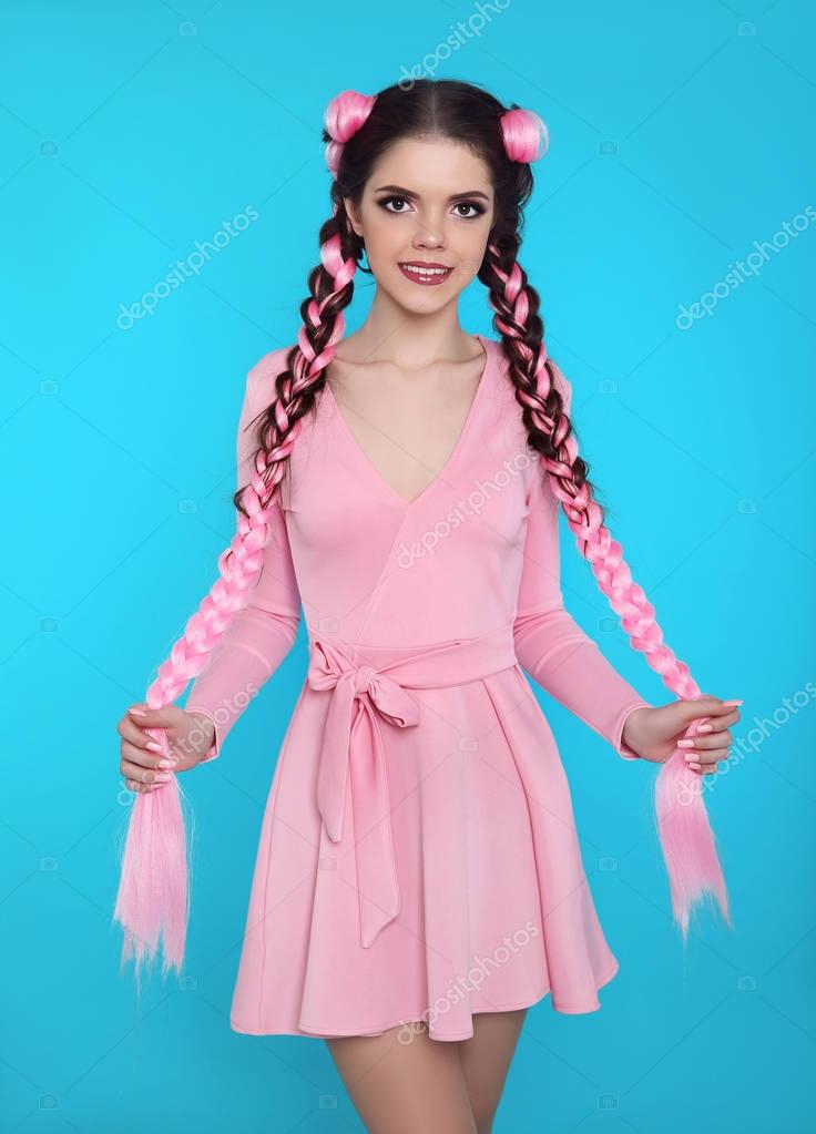 Pretty teen girl with two french braids from pink kanekalon, fashionable hairdo for youth, creative hairdresser beauty salon. Positive brunette posing in cute dress isolated on blue studio background.