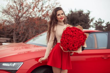 Çiçeklerle dolu güzel seksi bir kadın yeni kırmızı arabasıyla poz veriyor. Kırmızı etekli çekici esmer. Güzel sonbahar portresi. Sevgililer Günü. Romantik pahalı lüks hediye. 